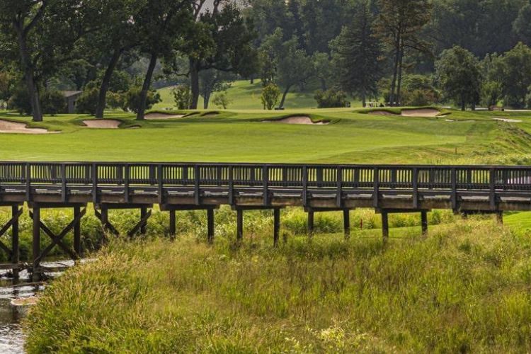 A wooden bridge leading to a lush fairway surrounded by trees and sand bunkers at Denver Country Club, a classic golfing destination.